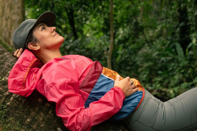 Woman in a brightly colored Cotopaxi brand rain jacket sitting on a tree branch.